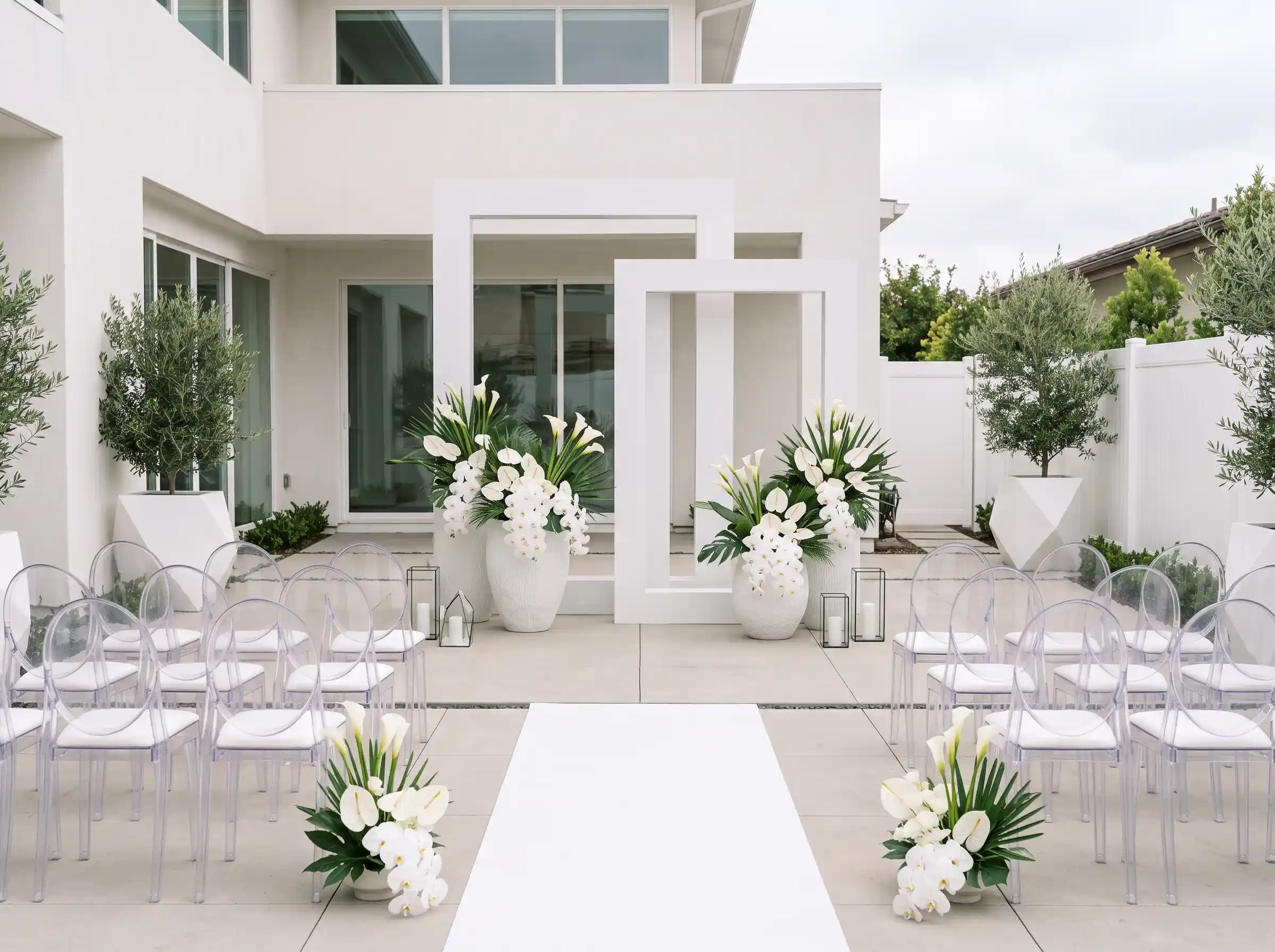 Modern minimalist backyard wedding ceremony with all-white color palette, ghost chairs, geometric white frame altar, white calla lily and orchid arrangements, white aisle runner, and olive trees against a contemporary home