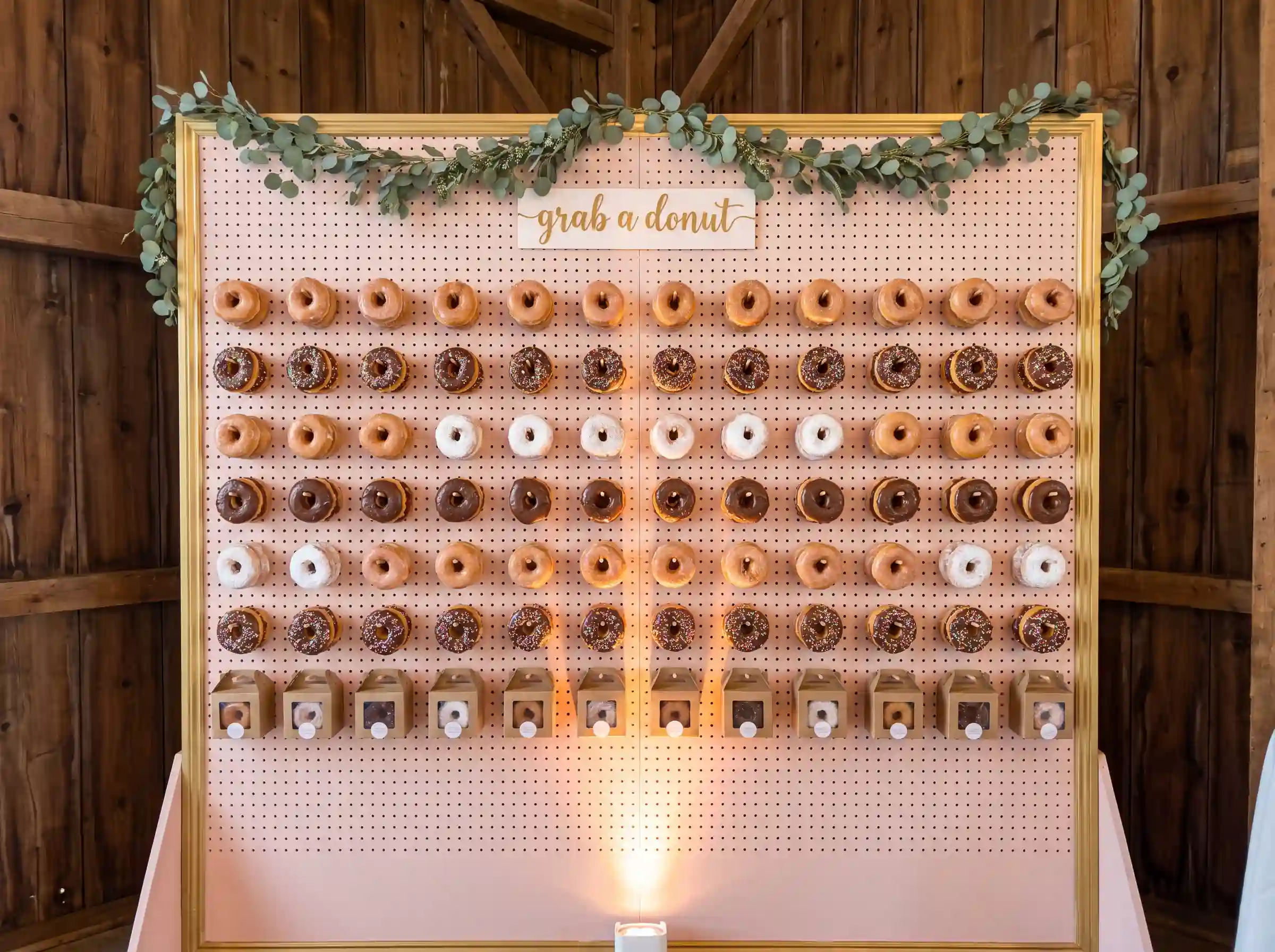 Blush pink pegboard donut wall with gold trim rows of glazed donuts eucalyptus garland and grab a donut sign
